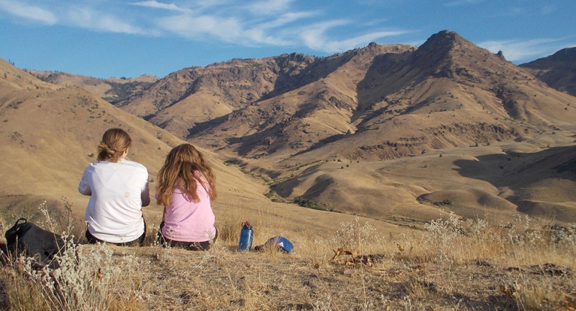 Two students sit facing away from the camera, looking out over a vast desert, mountainous landscape. 